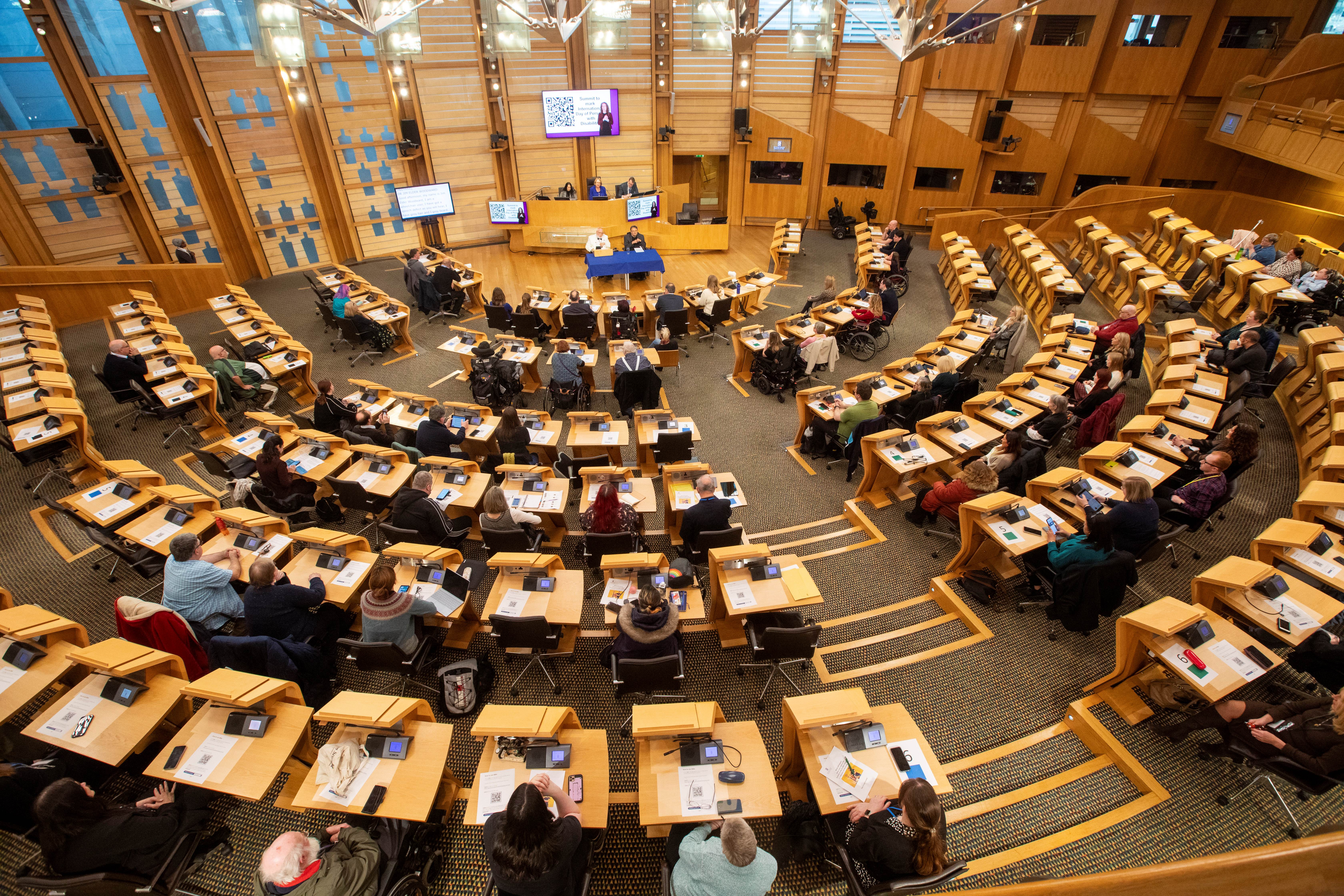 People sit at desks arranged in a semi-circle in the debating chamber of the Scottish Parliament. The audience includes several wheelchair users.